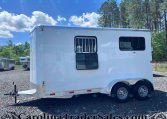 White horse trailer with two windows and a hitch, parked on gravel with trees and blue sky in the background.