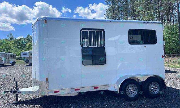 White horse trailer with two windows and a hitch, parked on gravel with trees and blue sky in the background.