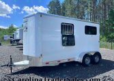 White horse trailer parked on a gravel lot with trees and a blue sky in the background; hitch and dual wheels visible on the right edge.