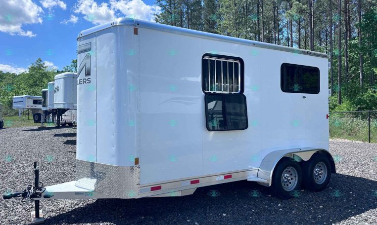 White horse trailer parked on a gravel lot with trees and a blue sky in the background; hitch and dual wheels visible on the right edge.