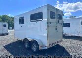 White enclosed horse trailer with side window and rear double doors, parked on a gravel lot under a bright blue sky with trees in the background.