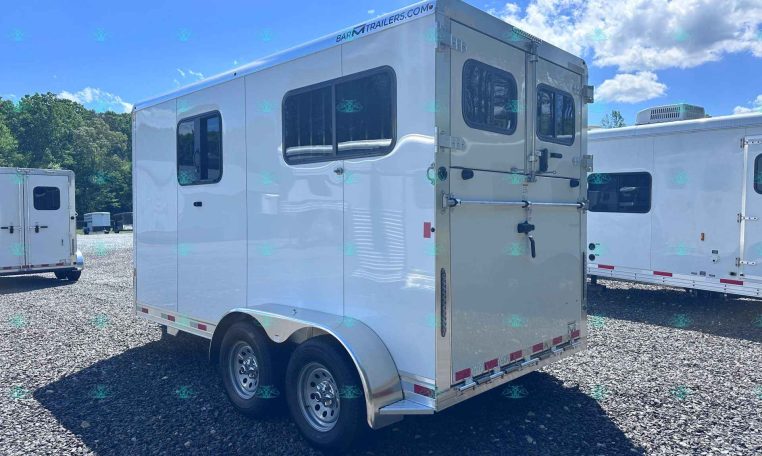 White enclosed horse trailer with side window and rear double doors, parked on a gravel lot under a bright blue sky with trees in the background.