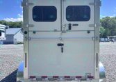 Rear view of a white double-door trailer with two small rounded windows, latches, and visible hinges in a gravel lot under a blue sky.
