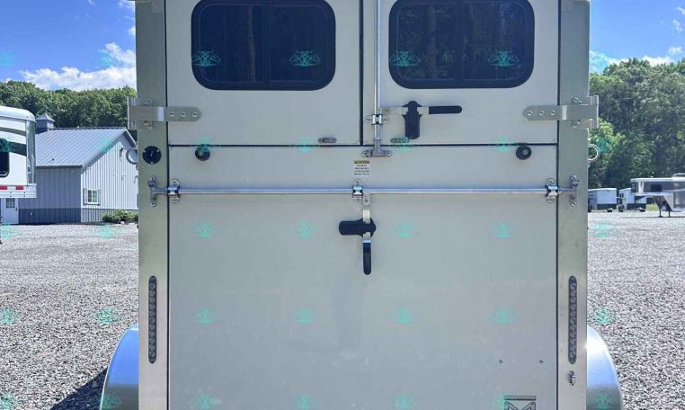 Rear view of a white double-door trailer with two small rounded windows, latches, and visible hinges in a gravel lot under a blue sky.
