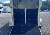 Open horse trailer with a lowered ramp, showing white and dark blue interior and a center divider in a gravel lot.
