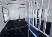 Interior of a horse trailer showing a metal divider with vertical bars and a cushioned saddle area on the left, windows on the side, and dark flooring for loading calm horses.
