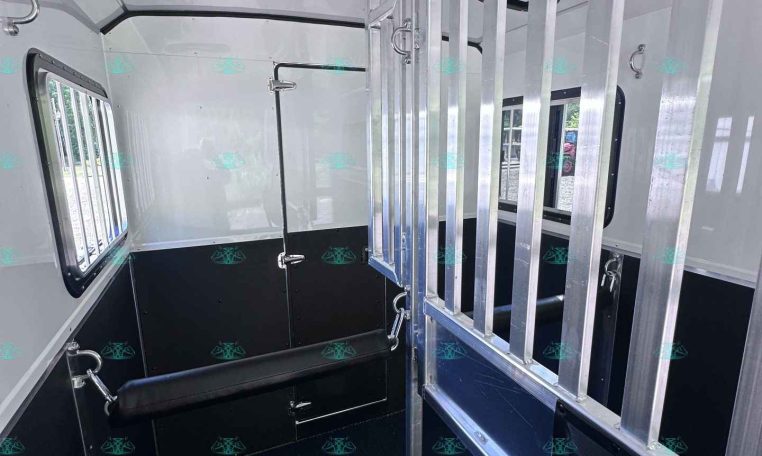 Interior of a horse trailer showing a metal divider with vertical bars and a cushioned saddle area on the left, windows on the side, and dark flooring for loading calm horses.