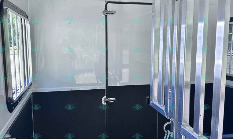 Interior of a horse trailer with white walls, a padded black bench, and metal divider rails along the doorway.