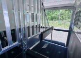 Interior view of a metal trailer with aluminum stairs and vertical railing, open side door and black non-slip floor, outdoor greenery visible outside.