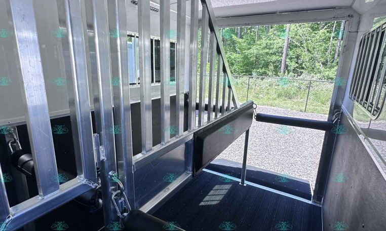 Interior view of a metal trailer with aluminum stairs and vertical railing, open side door and black non-slip floor, outdoor greenery visible outside.