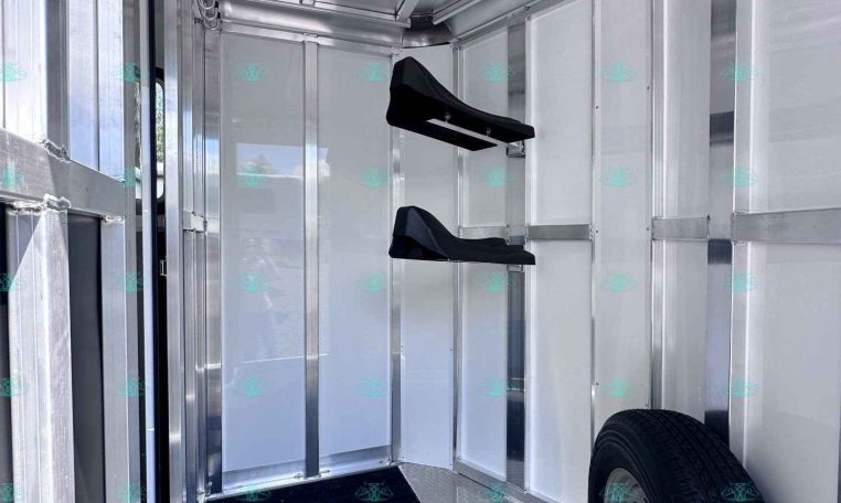 Interior of a aluminum trailered space showing a spare wheel, diamond-plate floor, and a pair of black fold-down saddle racks mounted to the wall, metal walls around.