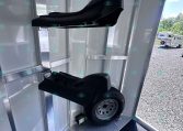 Two black saddle-style seats mounted inside a silver aluminum trailer, with a wheel resting on the diamond-plate floor nearby; clear exterior trailer lot visible through doorway.