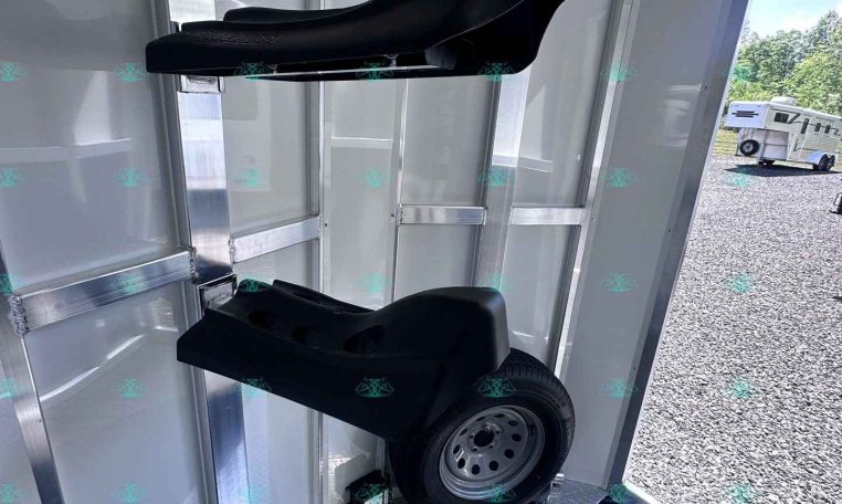 Two black saddle-style seats mounted inside a silver aluminum trailer, with a wheel resting on the diamond-plate floor nearby; clear exterior trailer lot visible through doorway.