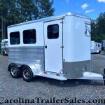 Silver aluminum horse trailer with two axles parked on a gravel lot, trees in the background.