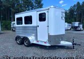 Silver aluminum horse trailer with two axles parked on a gravel lot, trees in the background.