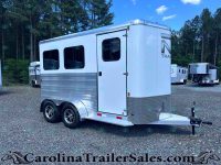 Silver aluminum horse trailer with two axles parked on a gravel lot, trees in the background.