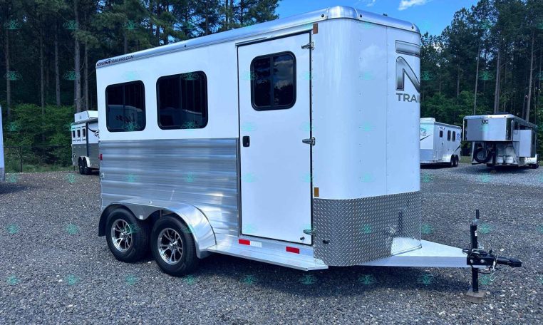 Silver aluminum horse trailer with two axles parked on a gravel lot, trees in the background.