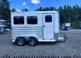 White aluminum horse trailer with two axles and rounded windows, parked on a gravel lot with trees in the background; watermark CarolinaTrailersSales.com across the bottom.