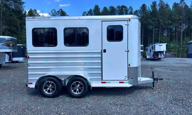 White aluminum horse trailer with two axles and rounded windows, parked on a gravel lot with trees in the background; watermark CarolinaTrailersSales.com across the bottom.