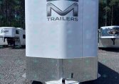 Front of a white enclosed trailer with a large chrome 'M' logo and the word TRAILERS, diamond-plate lower panel, parked on gravel.