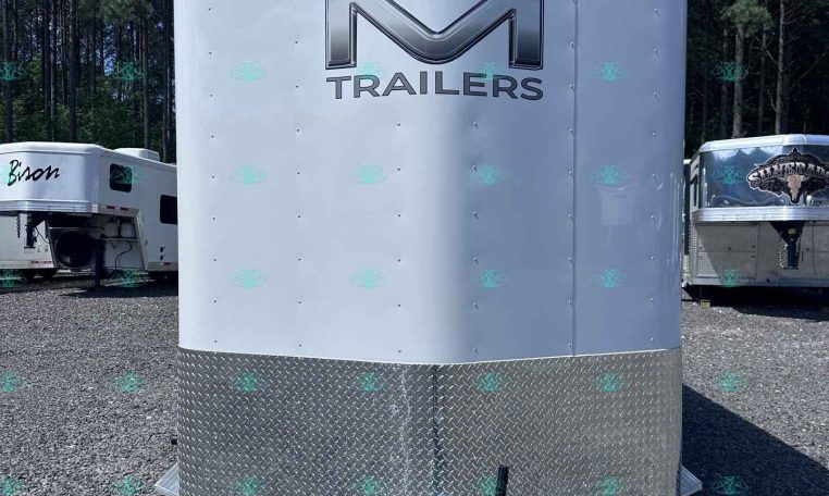Front of a white enclosed trailer with a large chrome 'M' logo and the word TRAILERS, diamond-plate lower panel, parked on gravel.