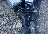 Close-up of a trailer tongue showing a black jack post, safety chains, electrical connector, and cables on a gravel surface. The RAM-branded label is visible on the jack.