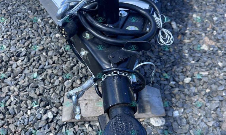 Close-up of a trailer tongue showing a black jack post, safety chains, electrical connector, and cables on a gravel surface. The RAM-branded label is visible on the jack.
