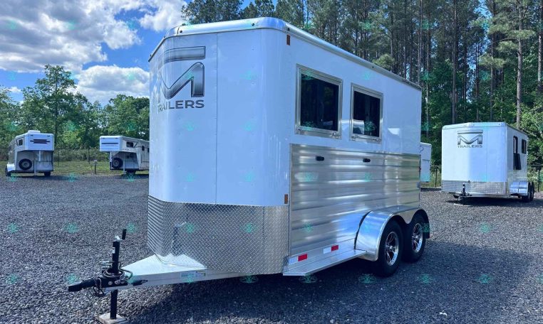Silver enclosed trailer on a gravel lot with other trailers and pine trees in the background, watermark reads CarolinaTrailersSales.com.