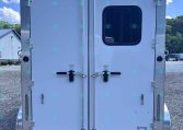 Rear view of an enclosed cargo trailer with double doors; a small rounded window is on the right door, multiple latches and hinges visible, and a reflective strip at the bottom on a gravel lot under a blue sky.