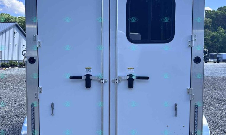 Rear view of an enclosed cargo trailer with double doors; a small rounded window is on the right door, multiple latches and hinges visible, and a reflective strip at the bottom on a gravel lot under a blue sky.