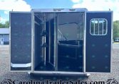 Open horse trailer with blue interior stalls and hooks on a gravel lot under a sunny sky.