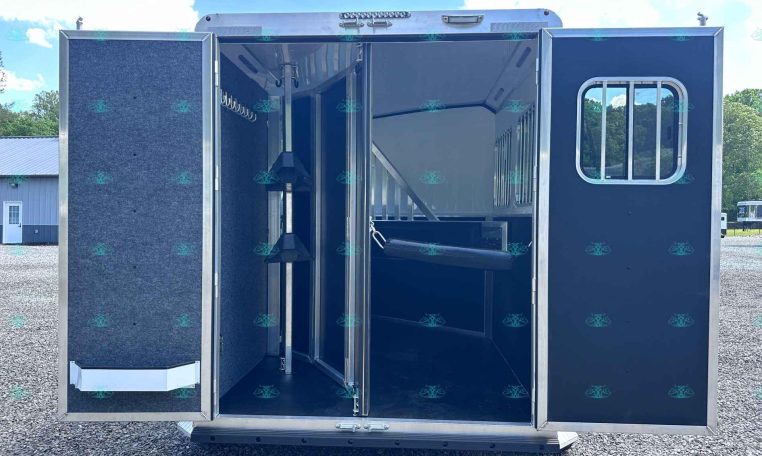 Open horse trailer with blue interior stalls and hooks on a gravel lot under a sunny sky.