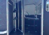 Interior of an empty horse trailer with partition stalls, metal rails, and rubber mat flooring, seen through open rear doors against a blue exterior.