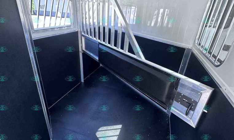 Interior of a horse trailer showing a metal divider, windows, and a black non-slip floor inside a partially partitioned stall.