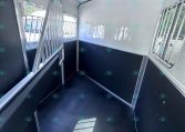 Interior of a white horse trailer with dark lower walls, metal divider rails creating separate stalls along each side, windows with bars on the right, and a narrow central aisle; sunlight streams in through the windows.