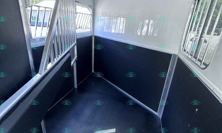 Interior of a white horse trailer with dark lower walls, metal divider rails creating separate stalls along each side, windows with bars on the right, and a narrow central aisle; sunlight streams in through the windows.