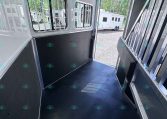 Inside a silver horse trailer with barred windows and black rubber flooring; another trailer is visible outside.