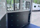 Interior of a white horse trailer showing two barred side windows, a black divider wall, and a tie ring; outside in a lot are other trailers.