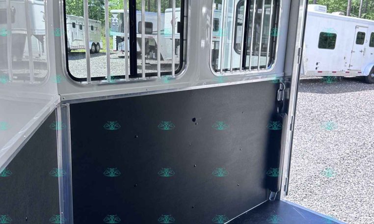Interior of a white horse trailer showing two barred side windows, a black divider wall, and a tie ring; outside in a lot are other trailers.