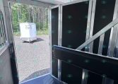 Interior of a trailer showing black partition walls with aluminum framing; open door reveals a white trailer outside on a gravel lot near trees.