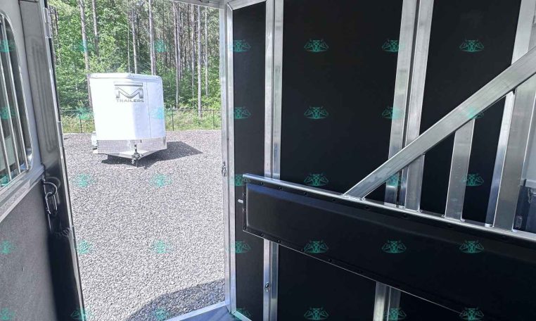 Interior of a trailer showing black partition walls with aluminum framing; open door reveals a white trailer outside on a gravel lot near trees.