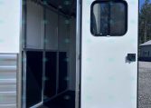 Open horse trailer door revealing empty interior stalls with metal partitions and overhead hooks.