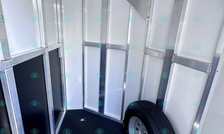 Interior of a silver aluminum cargo trailer with metal framing, wheel mounted in the corner and a spare tire visible on the floor.
