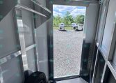 Inside a metal trailer toward an open back door; a spare wheel is visible on the floor and two white trailers are outside on a gravel lot with trees in the background.