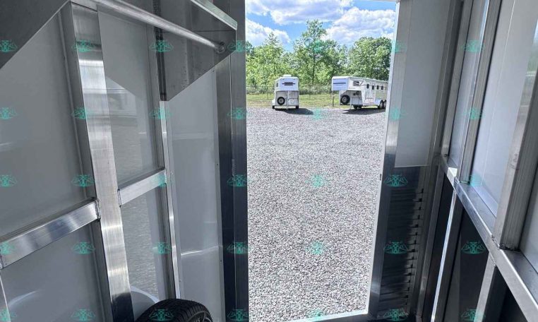 Inside a metal trailer toward an open back door; a spare wheel is visible on the floor and two white trailers are outside on a gravel lot with trees in the background.