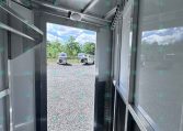 Interior of a metal trailer with the door open, looking out to a gravel lot and two white trailers in the distance under a blue sky.