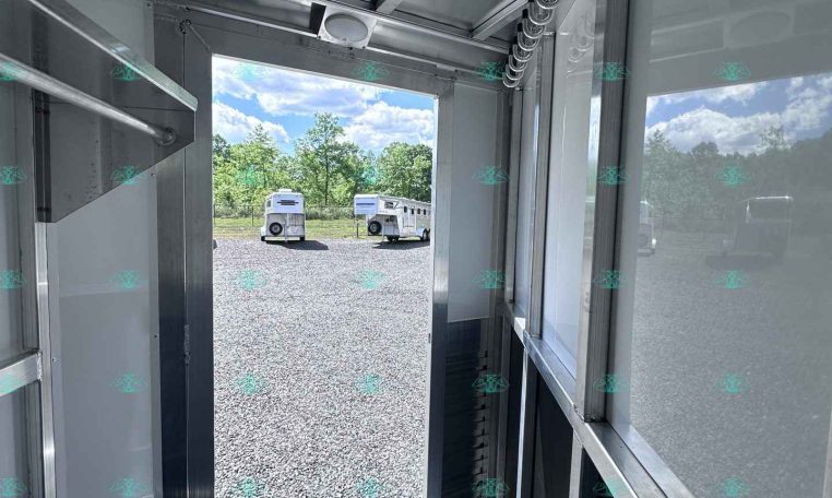 Interior of a metal trailer with the door open, looking out to a gravel lot and two white trailers in the distance under a blue sky.