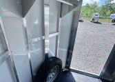 Interior of a silver trailer showing a mounted spare wheel and metal framing; open door reveals a gravel lot with horse trailers outside.