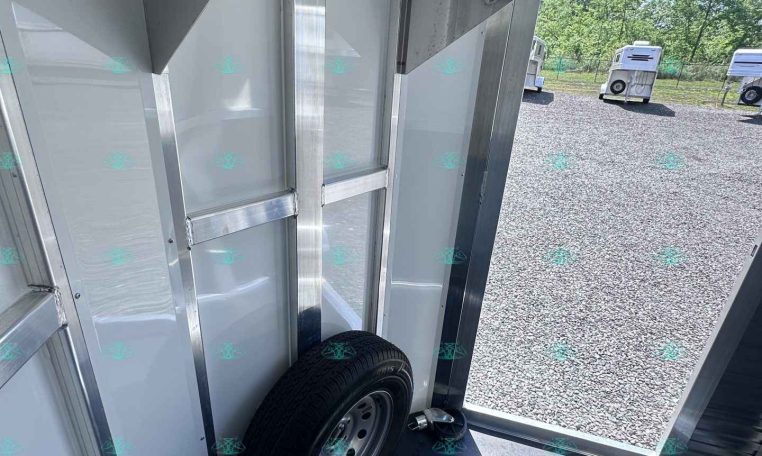 Interior of a silver trailer showing a mounted spare wheel and metal framing; open door reveals a gravel lot with horse trailers outside.