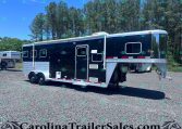 Black and silver horse trailer on a gravel lot with a wooded backdrop and blue sky behind it.